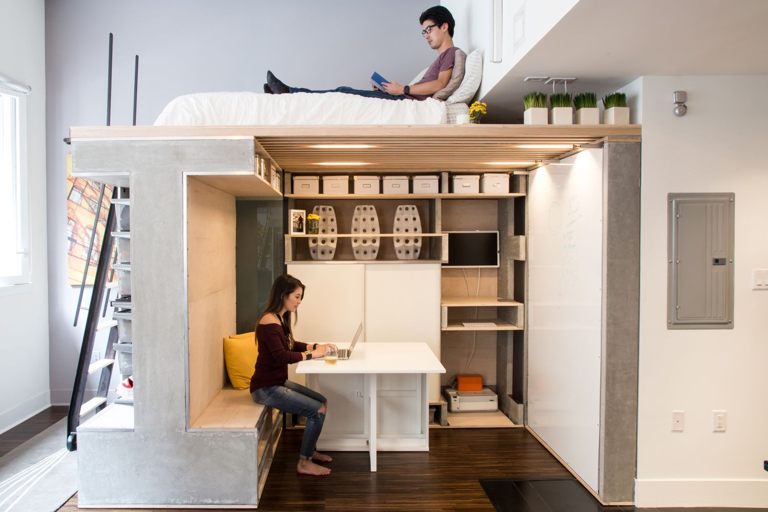 Studio avec espace de vie en loft où l'homme lit en haut et espace de travail en bas où la femme travaille sur un ordinateur portable.