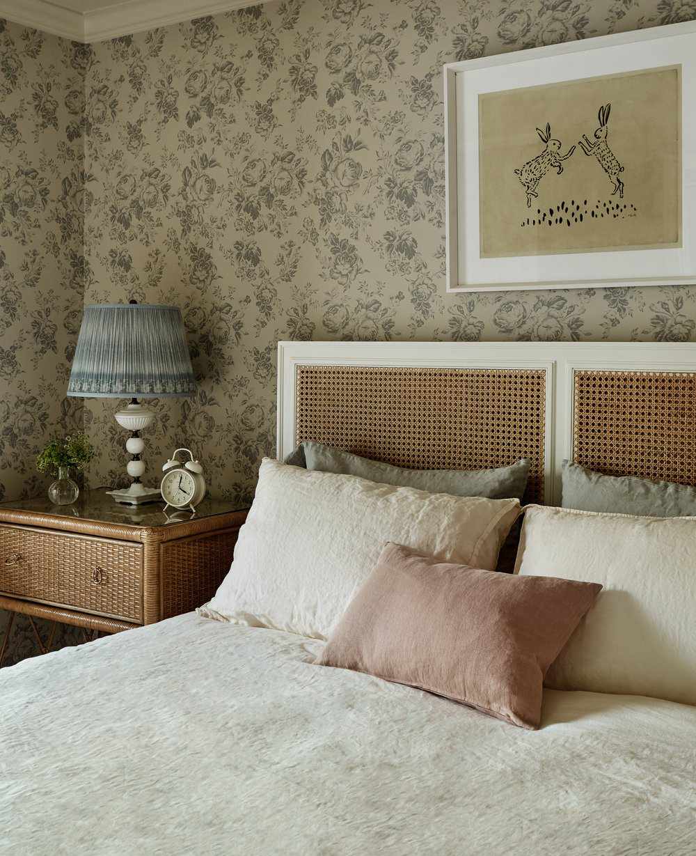 Une chambre à coucher remplie de papier peint floral, de meubles en rotin tressé, d'un abat-jour plissé et d'une literie en lin souple.