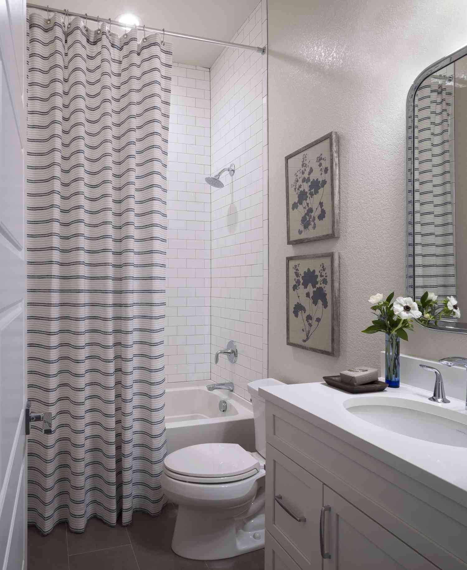 Une salle de bain blanche a un rideau de douche rayé et deux images beiges avec des fleurs bleues assorties au rideau rayé.