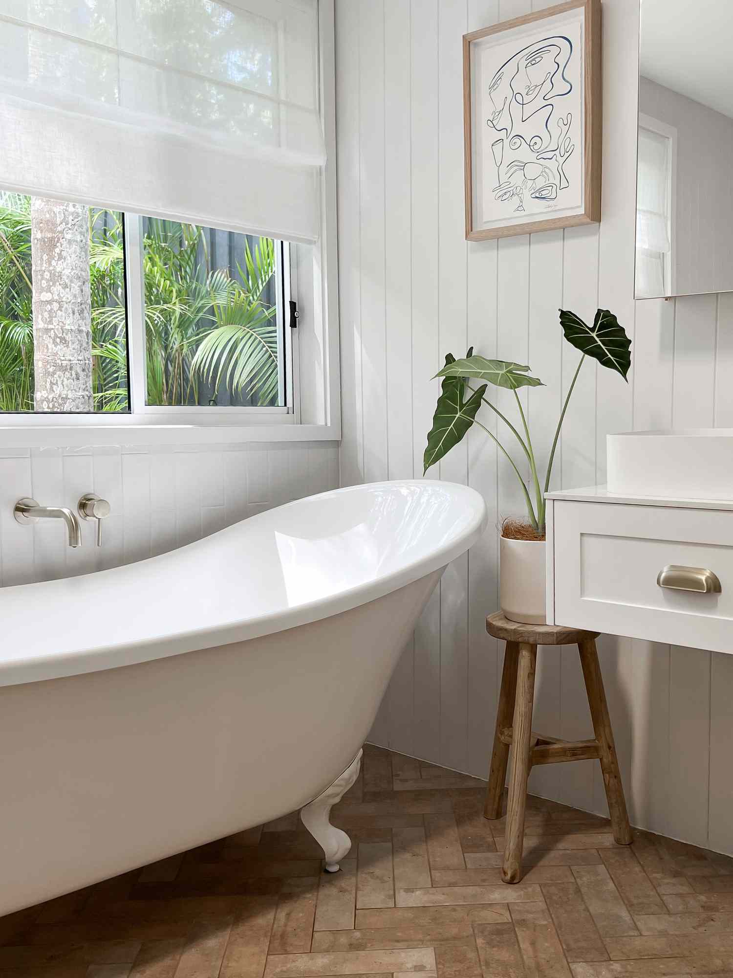 Une salle de bains recouverte de shiplap a des accents de bois simples comme un tabouret tenant une plante d'intérieur près de la grande baignoire.