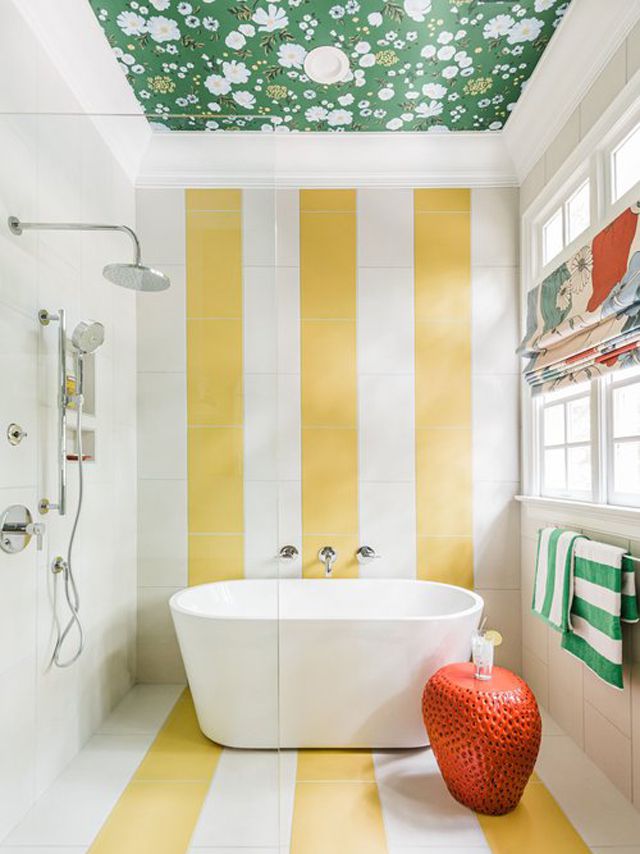 Salle de bain rayée jaune et blanche avec baignoire autoportante, plafond en papier peint floral vert et tabouret en céramique orange.