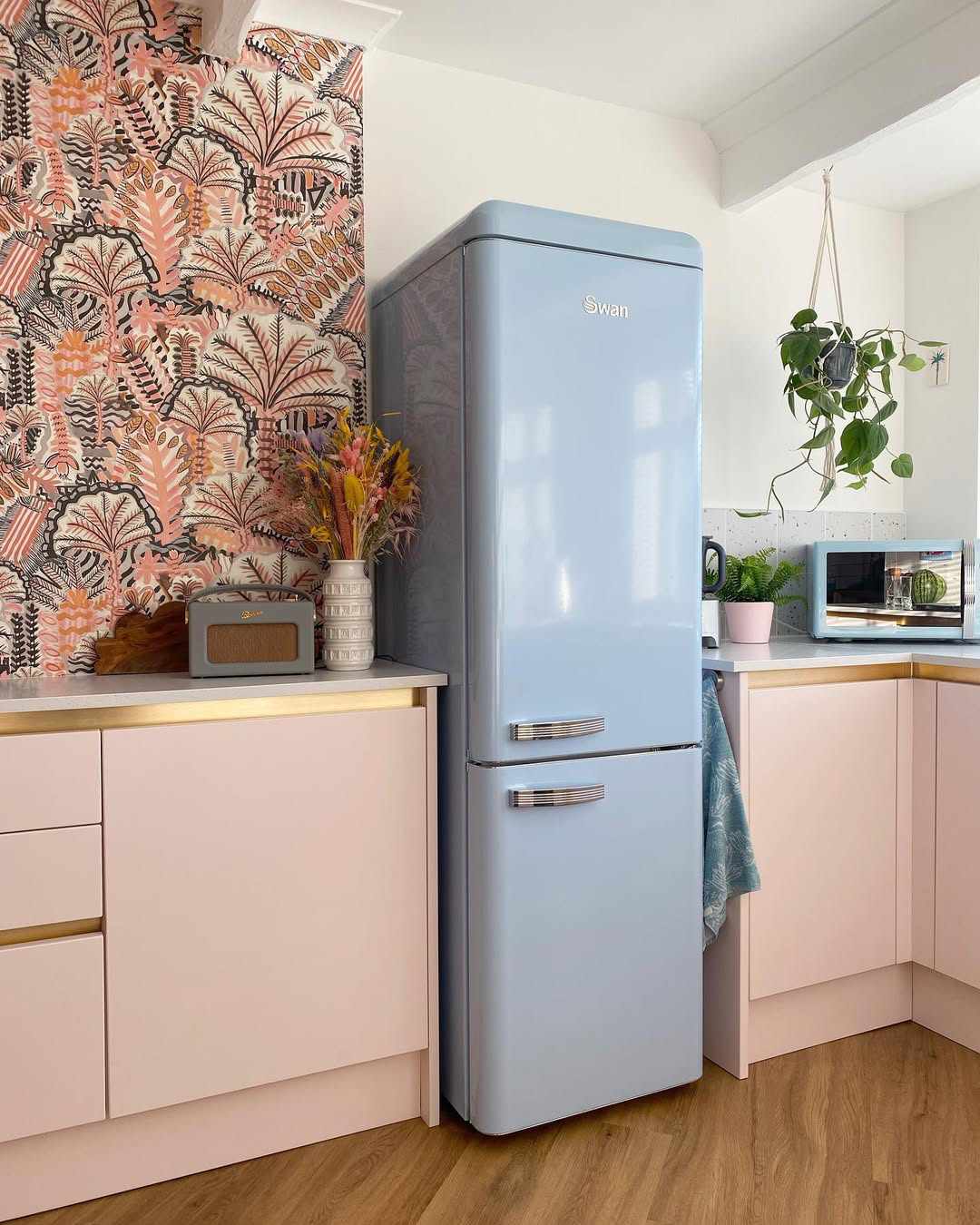Une cuisine pastel avec des meubles roses, un papier peint aux accents floraux et un réfrigérateur bleu clair de Swan.